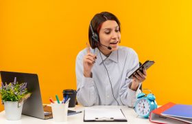 impressed young call center girl wearing headset sitting desk holding looking mobile phone with raised finger isolated orange 280x180